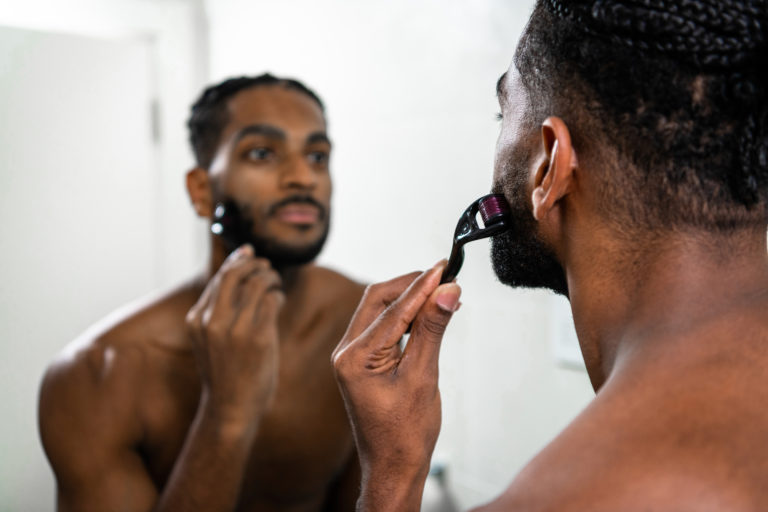 Young man rolling a derma roller on his bearded face in front of a mirror in the bathroom.