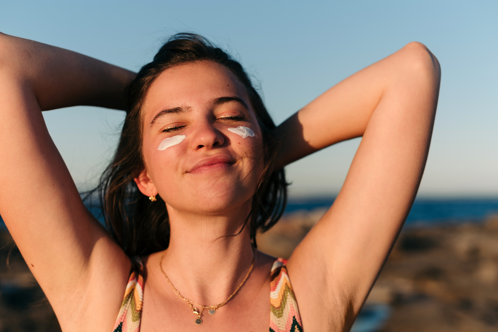 Young woman outdoors with sunscreen on her cheeks, closing her eyes and smiling.