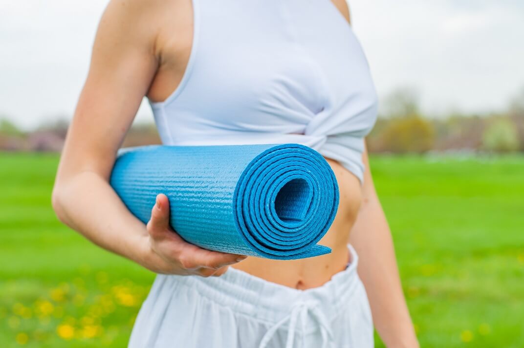 Woman standing in a field holding a folded up yoga mat