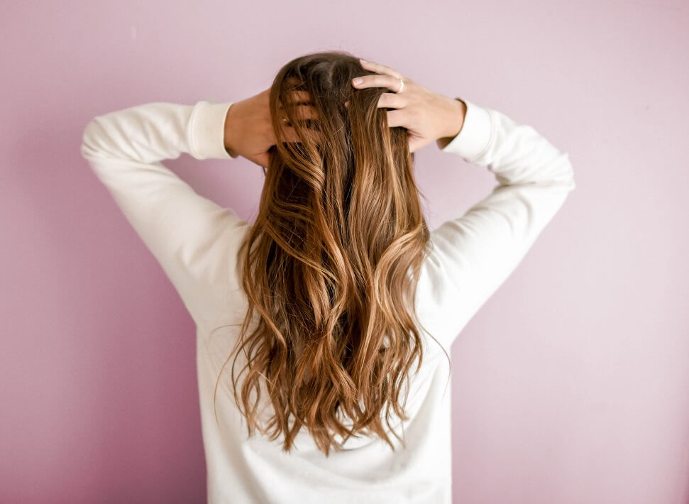 Femme debout avec son visage vers un mur rose et ses mains dans ses longs cheveux